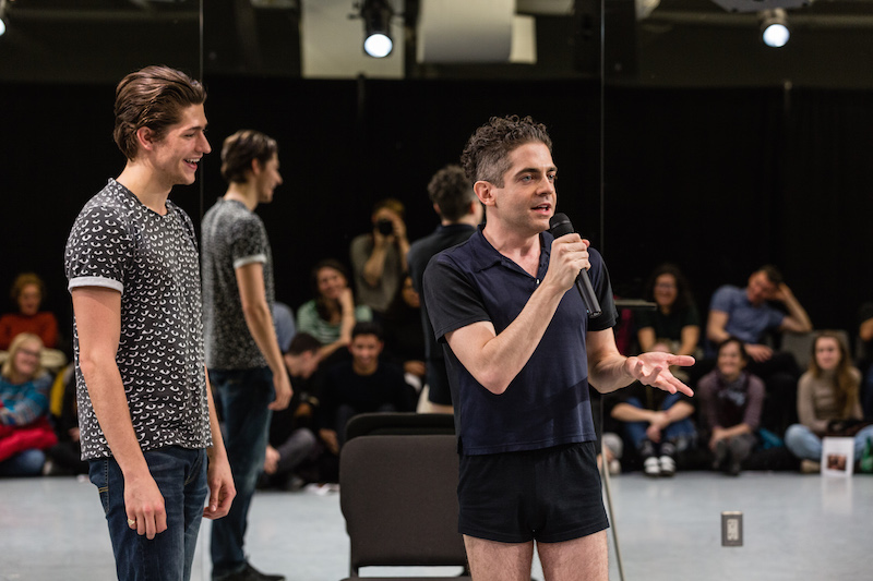 Jack Ferver in short black shorts talks to the audience while another man stands next to him while laughing